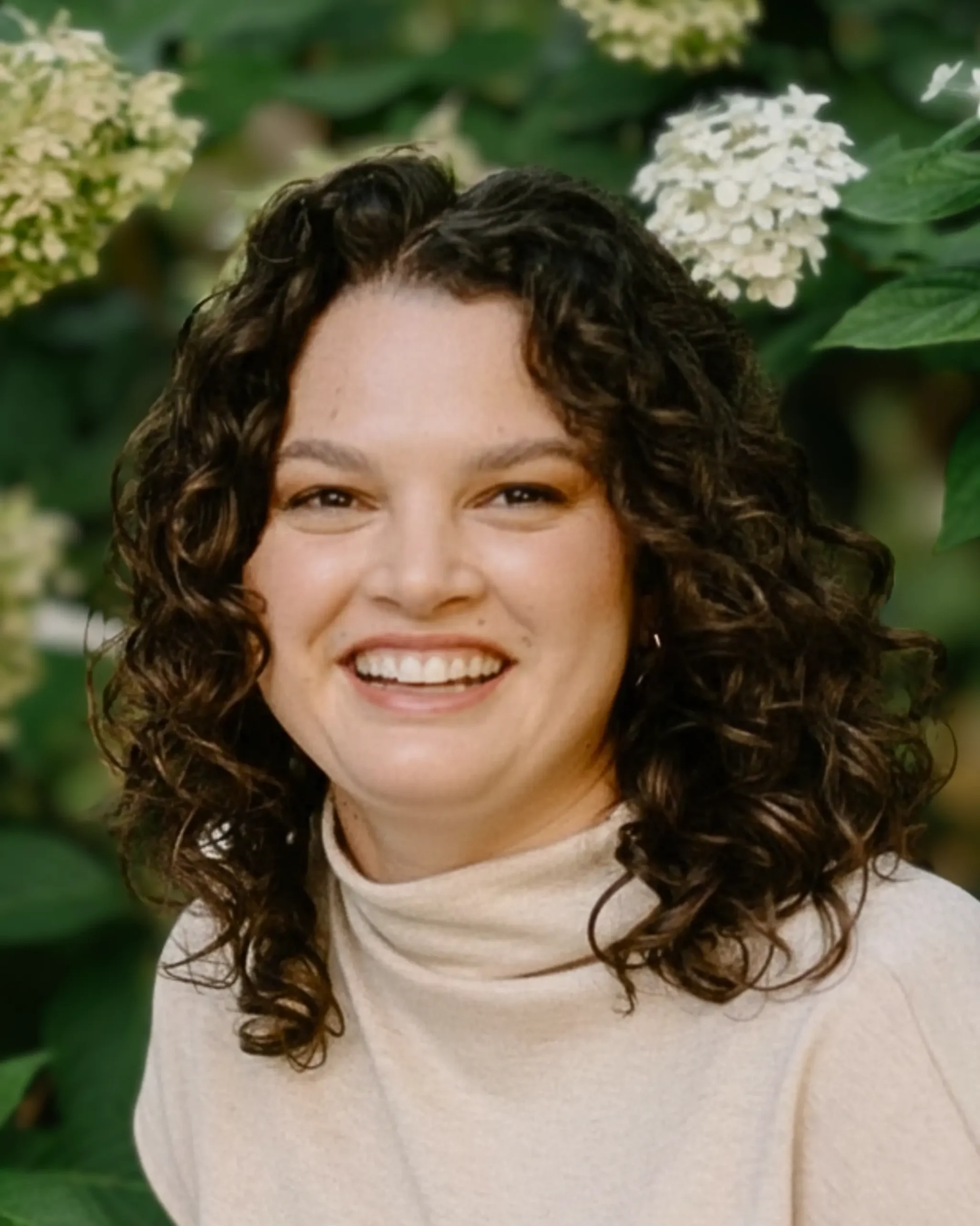 Smiling woman with shoulder-length dark curly hair wearing a light beige turtleneck, photographed outdoors in front of green foliage and white flowers.