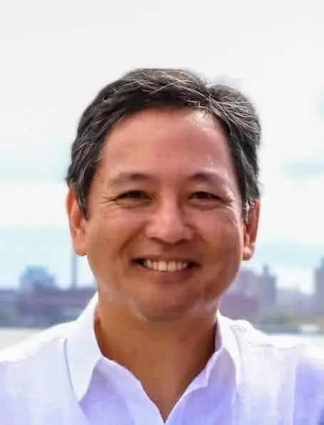 Smiling man with short salt-and-pepper hair wearing a white button-down shirt, photographed outdoors with a city skyline in the background.